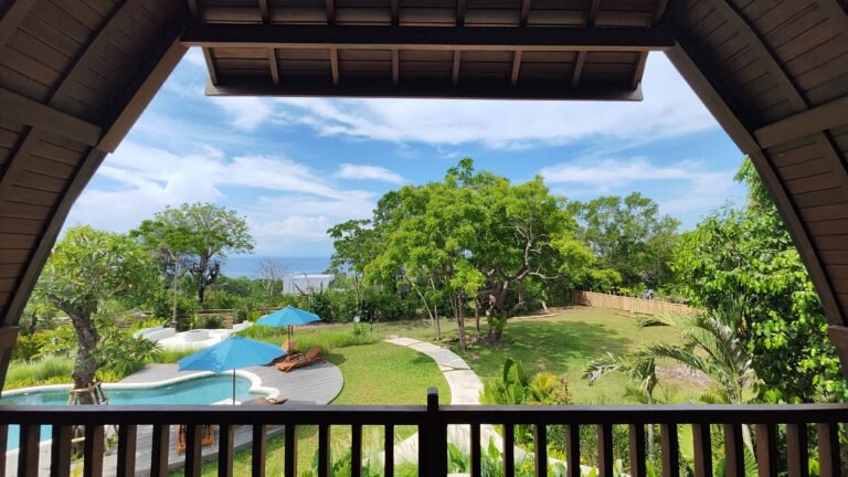 View from The Mahaloka balcony, framed by the roofline, overlooking the pool, large trees, gardens, and distant ocean horizon.