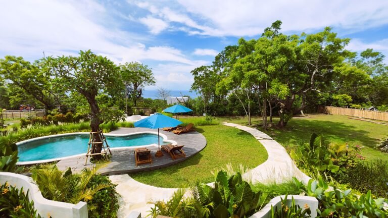 Vibrant high-angle panoramic view from The Mahaloka balcony showing the pool, deck, gardens, trees, and ocean under a blue sky.