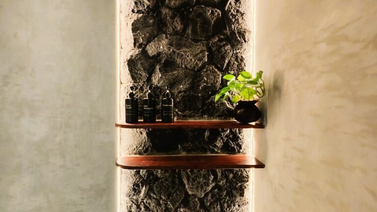 Detail shot of premium bathroom amenities on wooden shelves against a backlit natural stone feature wall in The Mahaloka bathroom.
