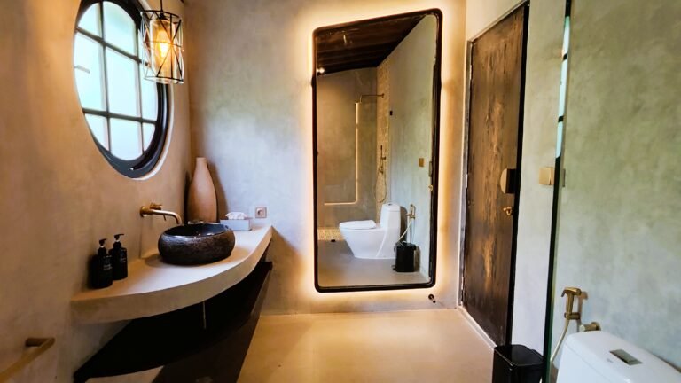 Angled view of The Mahaloka bathroom showing the backlit mirror reflecting the shower, stone sink, round window, and concrete walls.