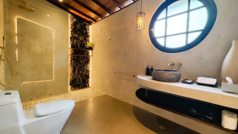The Mahaloka bathroom sanctuary featuring a natural stone sink, round window, polished concrete vanity, and rainfall shower reflection.