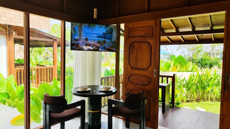 View of the intimate dining table and chairs in The Mahaloka lumbung living area, with TV mounted and view to the garden deck.