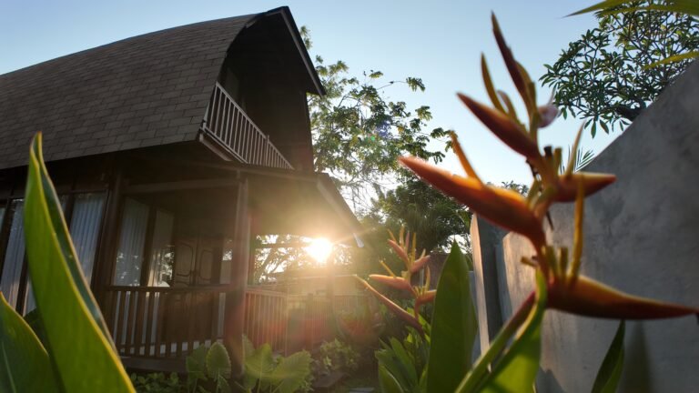 View of The Mahaloka lumbung villa exterior at sunset with sun flare filtering through a tropical orange flower in the foreground.