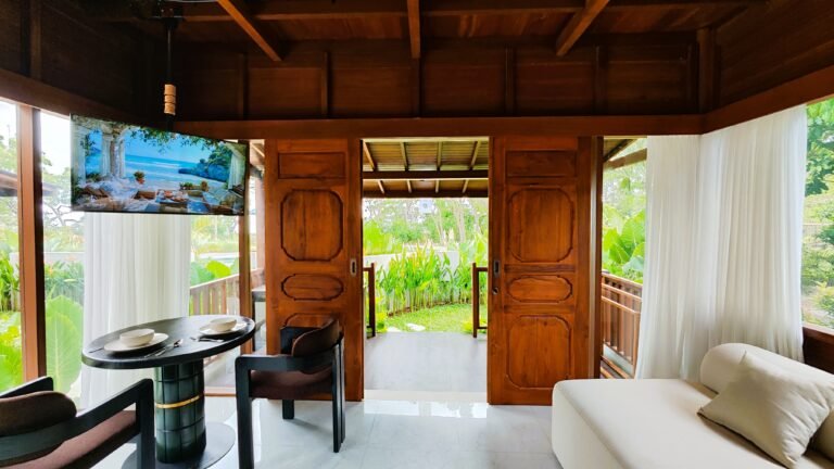 Wide view of The Mahaloka lumbung living area showing the sofa, dining table, and large wooden doors opening to the garden deck.