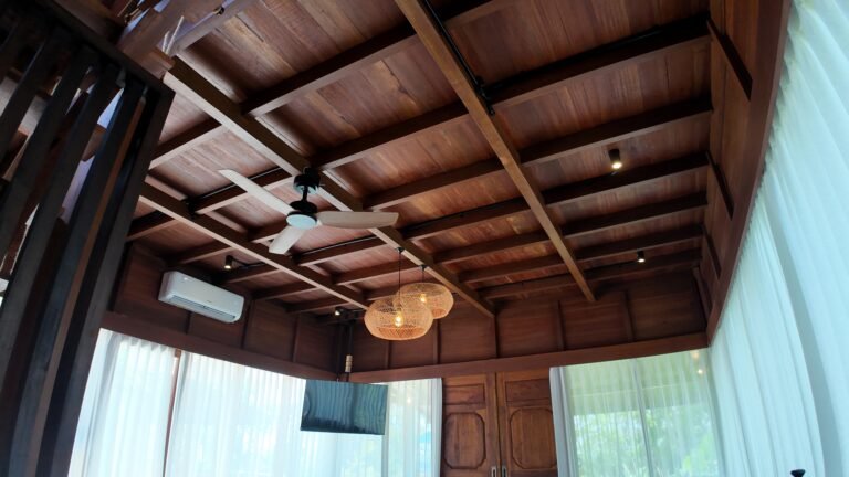 Looking up at the rich wooden beamed ceiling, fan, AC unit, and woven pendant lights in The Mahaloka living area.
