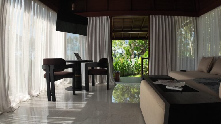 Interior view of The Mahaloka lumbung living room showing the sofa, coffee table, and dining set, looking towards the open deck door.