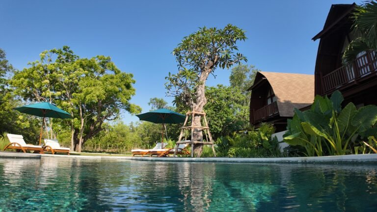 Low angle view across the sparkling shared pool towards The Mahaloka's lumbung villas and lush gardens under a blue sky.